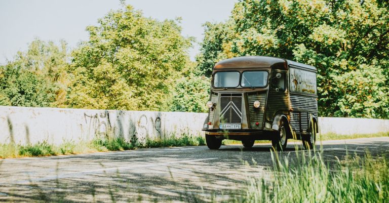 A vintage black delivery truck driving along a road with green trees and a concrete barrier in the background.