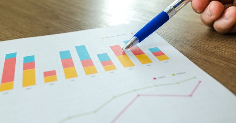 Close-up of a hand holding a blue pen pointing at a colorful bar chart on paper, on a wooden table.