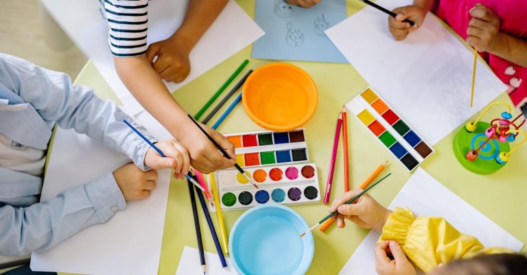 Children with various skin tones painting with watercolors and brushes at a colorful table, surrounded by art supplies and paper.