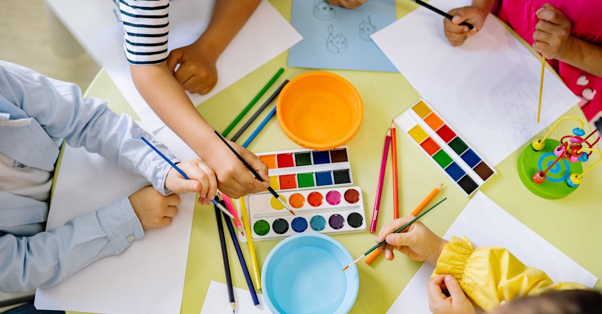 Children with various skin tones painting with watercolors and brushes at a colorful table, surrounded by art supplies and paper.