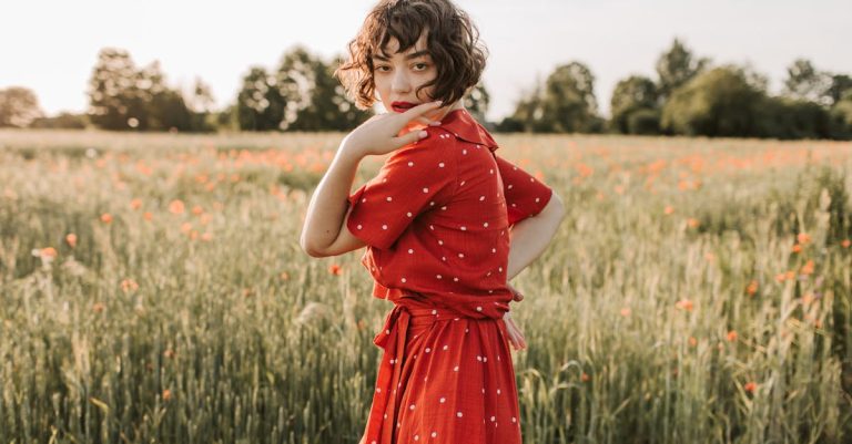 A woman with curly hair standing in a field of tall grass and flowers, wearing a red polka dot dress.
