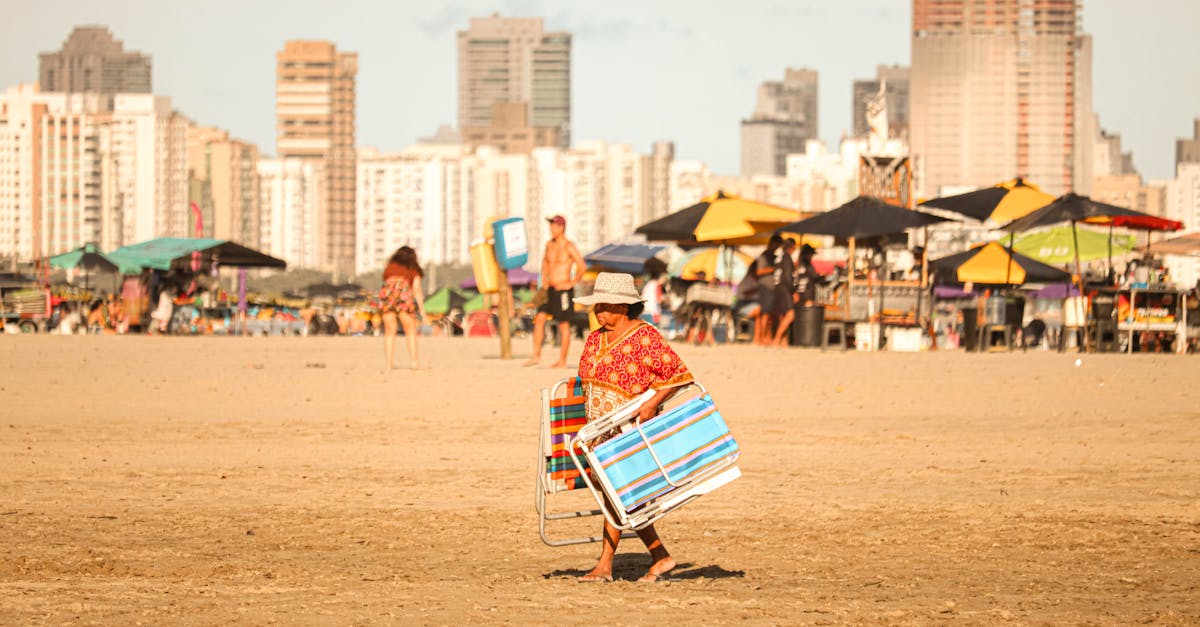 An elderly woman carrying a folded beach chair on a sandy beach with high-rise buildings in the background.