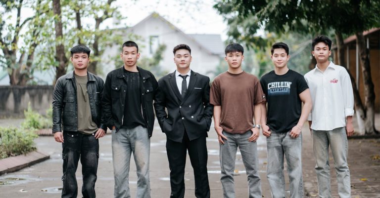 A group of six men standing side by side on a street, dressed in casual and smart casual outfits, with a blurred background of trees and buildings.