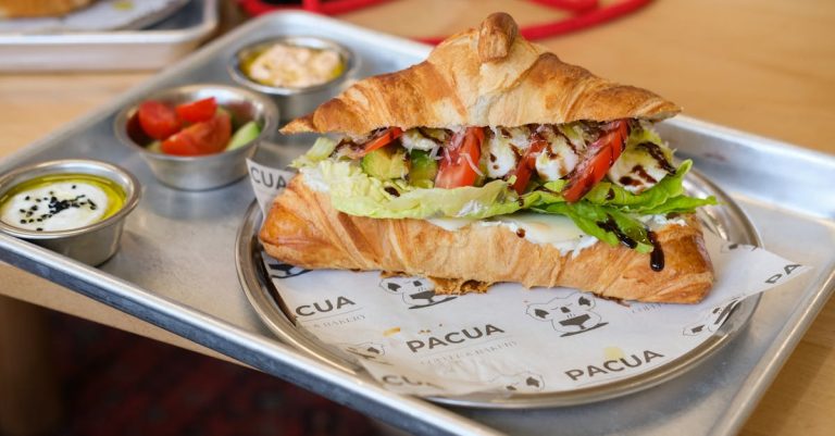 A croissant sandwich with lettuce, tomato, and other vegetables, accompanied by small containers of sauces and a side salad.