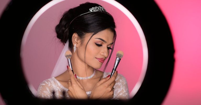 A bride's face and upper body with radiant, dewy skin, wearing a tiara, earrings, and necklace, holding makeup brushes.